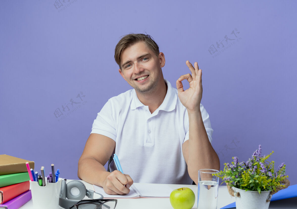 书桌面带微笑的年轻帅哥坐在书桌旁 拿着学习用具 摆出一副很好的姿势 在蓝色的笔记本上写着什么工具男性年轻
