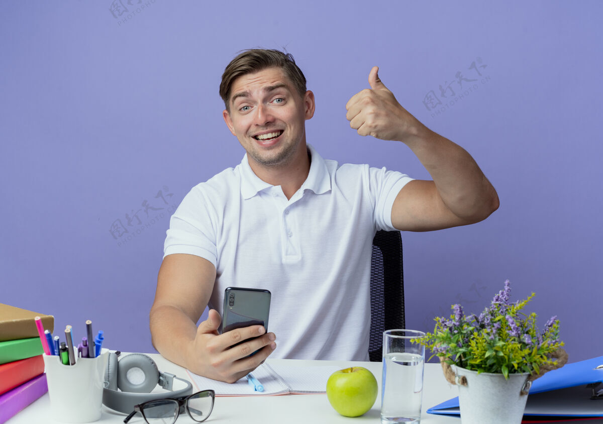 学生面带微笑的年轻英俊的男学生坐在书桌旁 手里拿着学习工具 举着电话 他的大拇指孤零零地竖在蓝色的桌子上拇指书桌电话
