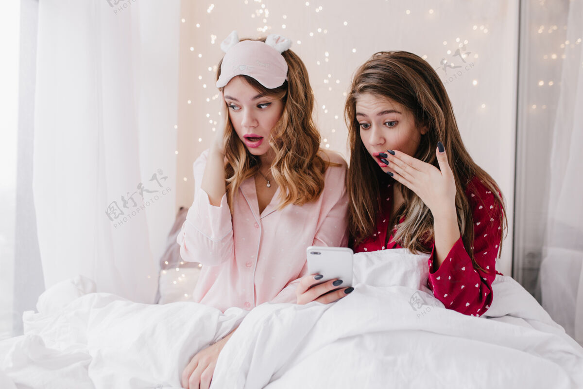 室内震惊的长发女士坐在白色毛毯下发短信无聊的金发女孩穿着粉色睡衣的室内照片睡眠面罩成人毛毯