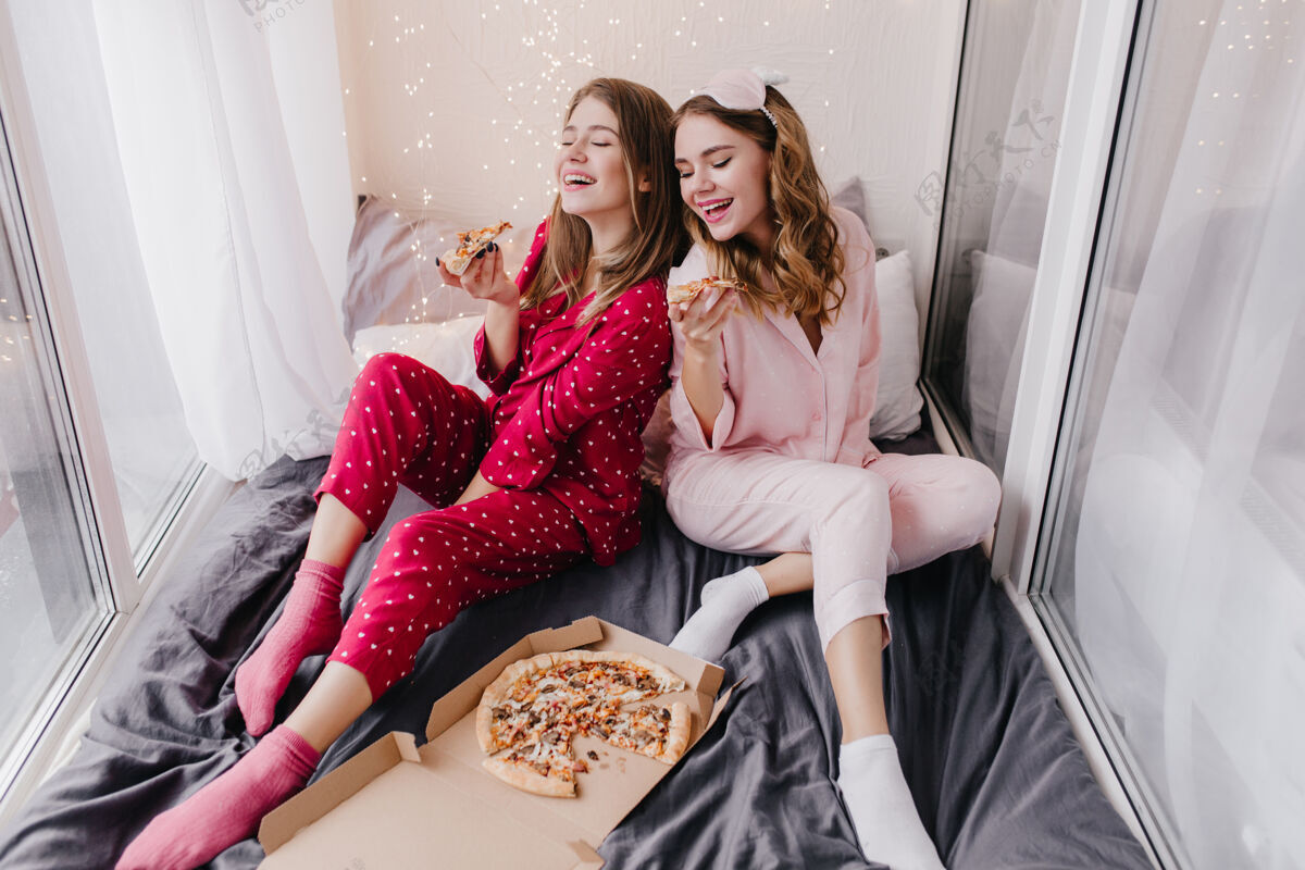 午餐穿着粉色袜子的女孩和最好的朋友一起吃比萨饼两个穿着睡衣的姐妹在床上享受意大利美食的室内照片漂亮金发床