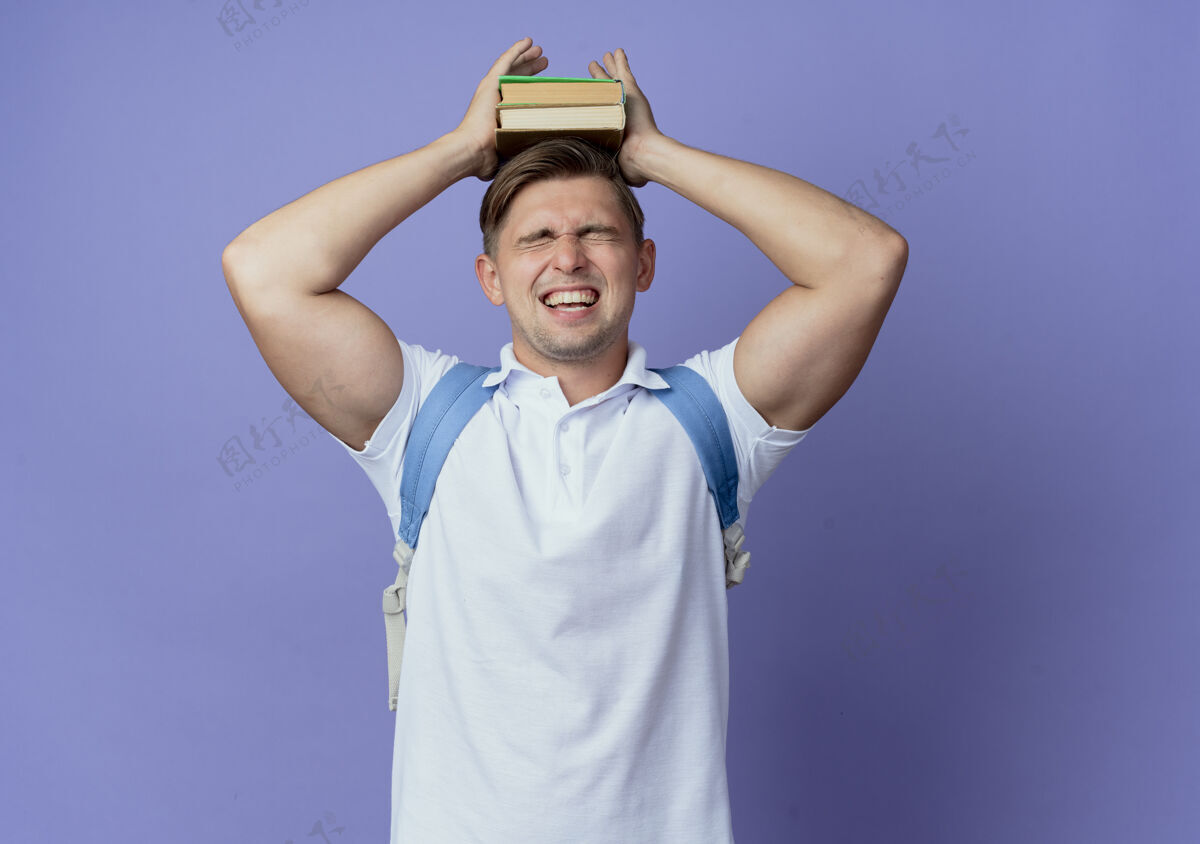 穿闭上眼睛快乐的年轻英俊的男学生戴着背包把书放在头上孤立在蓝色男性背部帅气