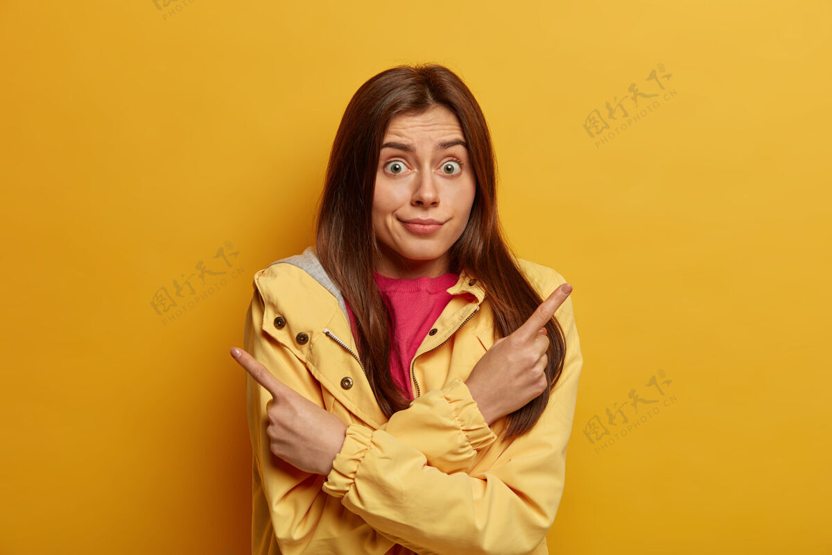 促销不知道犹豫的黑头发女人的照片指向一边 在两个选项中选择 有惊讶的表情 穿着夹克 对着黄色的墙壁摆姿势 说最好看看产品室内选择