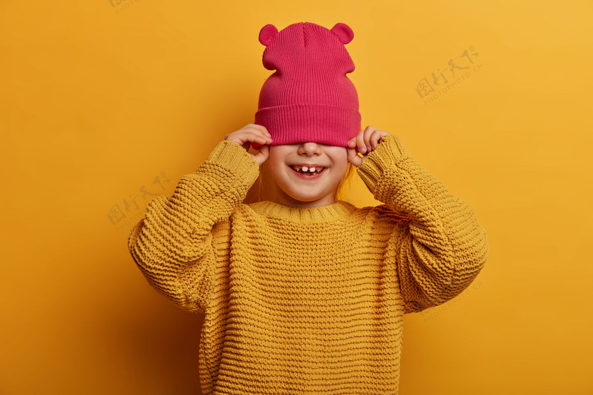满意孩子 快乐 幸福的概念无忧无虑的顽皮女孩用帽子遮住半张脸 试图躲着别人 穿着宽松的针织毛衣 孤立在黄色的墙上 得到了美丽的微笑欢笑请积极
