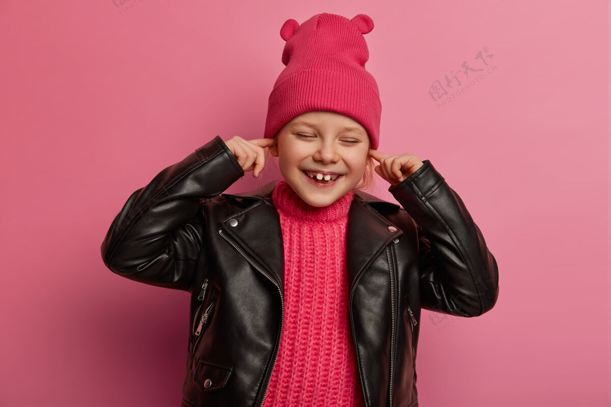 罗西快乐的小学龄前儿童捂着耳朵 把食指放在耳孔里 避免听到大声的音乐 有快乐的表情 戴着带耳朵的粉红色帽子和皮夹克 不想听噪音大声孩子咯咯笑