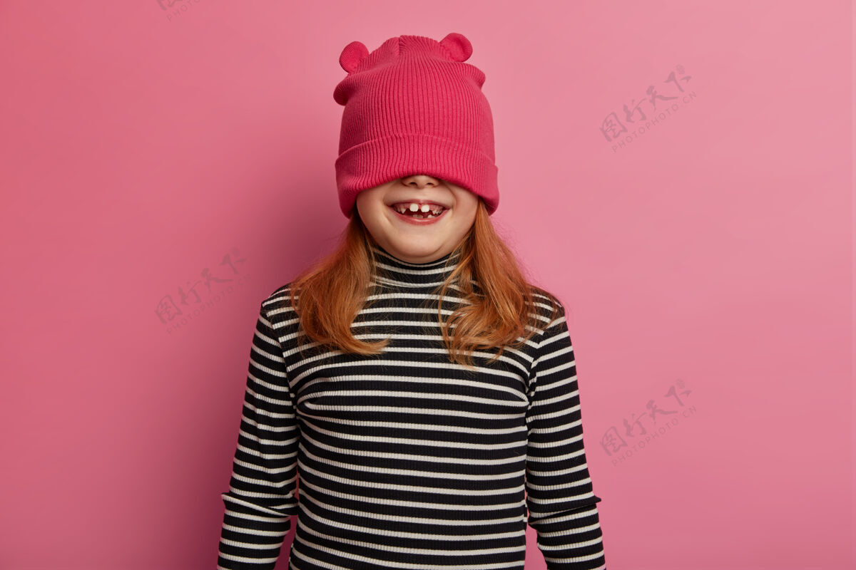 条纹顽皮的姜黄色小女孩的水平镜头用帽子遮住眼睛 穿着条纹套头衫 有一张滑稽的脸 在粉色粉彩墙上摆姿势 躲在帽子下面 和朋友或父母一起玩情绪粉彩感觉
