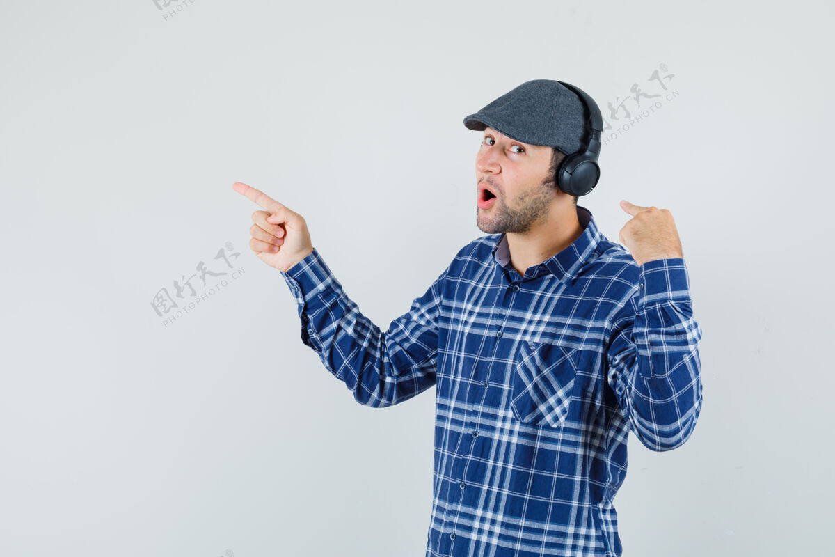 自信年轻人指着他的耳机 站在一边 穿着衬衫 戴着帽子 从正面看去前面胡须耳机
