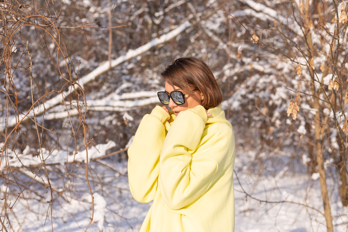 年轻阳光明媚的日子里 雪景冬林中一位年轻美女的画像 身着黄色大套头衫 戴着墨镜 享受着阳光和雪帽子自然夹克