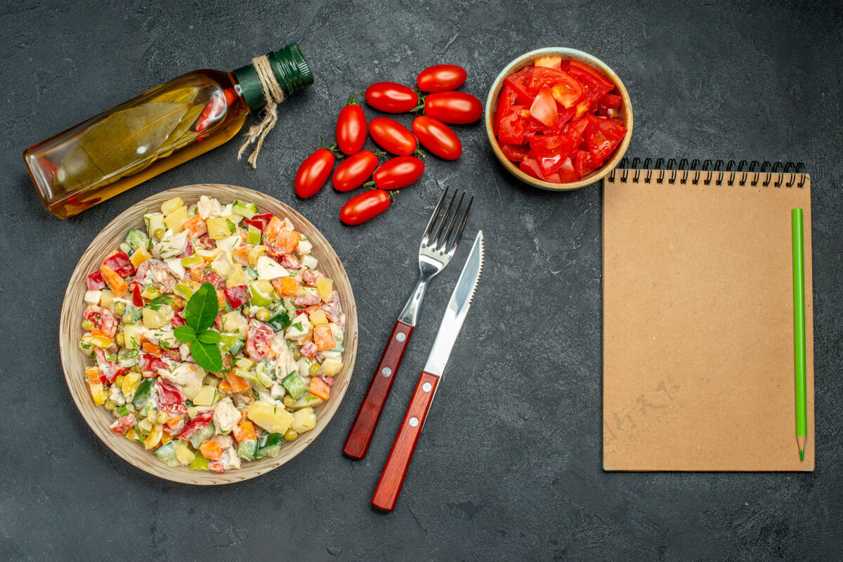 午餐蔬菜沙拉的顶视图 深灰色背景上有西红柿 油瓶餐具和记事本盘子记事本背景