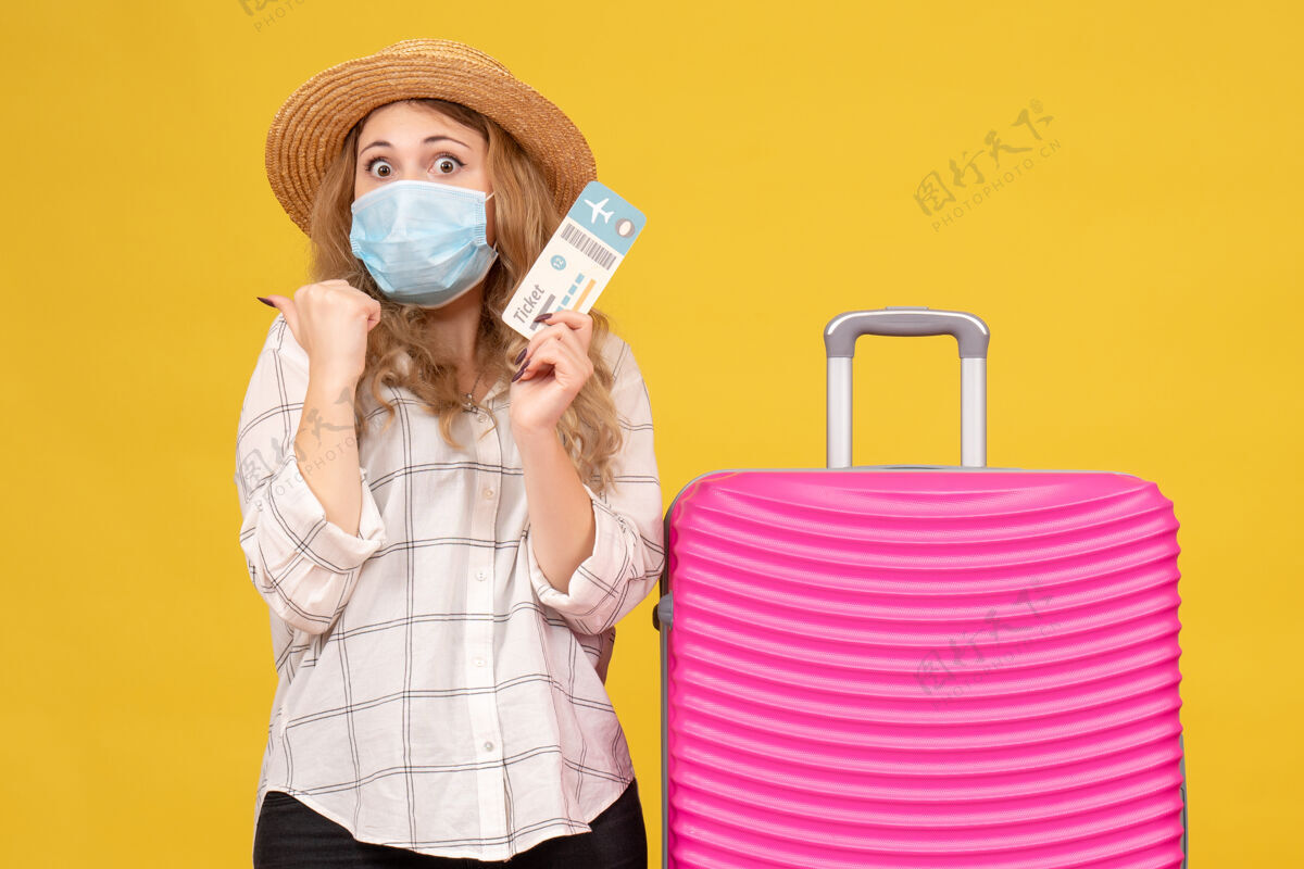 药前视图惊讶的旅行女孩戴着面具 出示车票 站在黄色的粉红色包附近站制服包