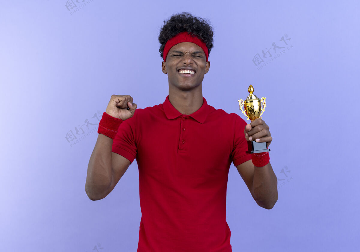 手势年轻的美国黑人运动男子戴着头带和腕带 紧闭双眼 拿着优胜者杯 在蓝色的舞台上表现出“是”的姿态蓝色杯子冠军