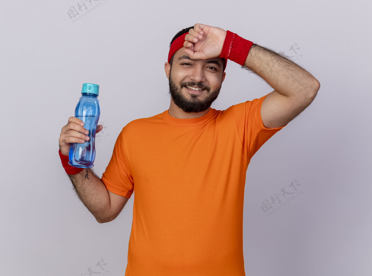 头带微笑着的年轻人戴着头带和腕带拿着水瓶 用手擦额头 在白色背景上隔离湿巾前额抱着