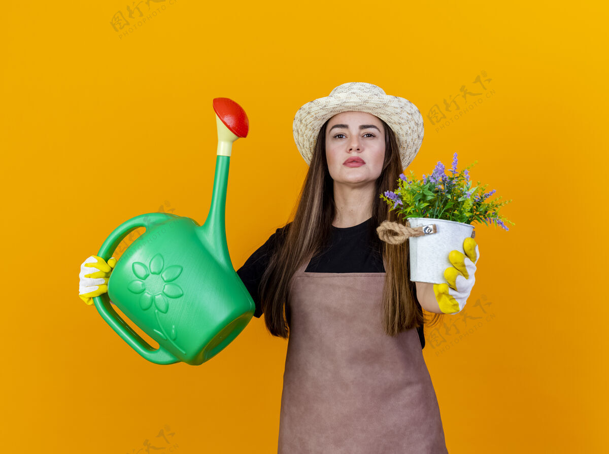 花自信美丽的园丁女孩穿着制服 戴着园艺帽 戴着手套 举着浇灌罐 拿着花盆里的花 对着隔离在橙色背景下的摄像机信心制服帽子