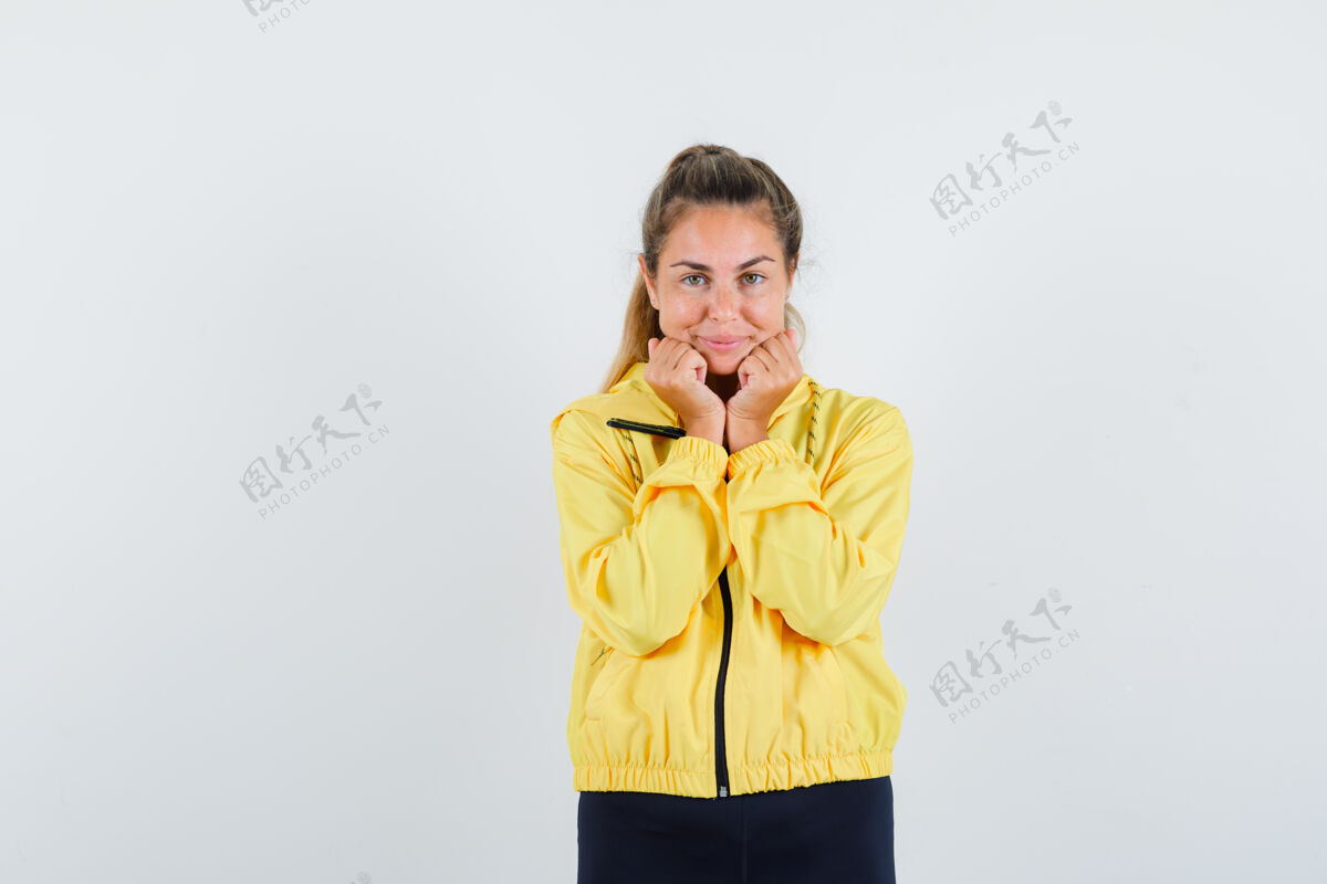 漂亮穿着黄色棒球服和黑色裤子的金发女人握紧拳头 把手放在下巴下面 看起来很开心美女模特夹克