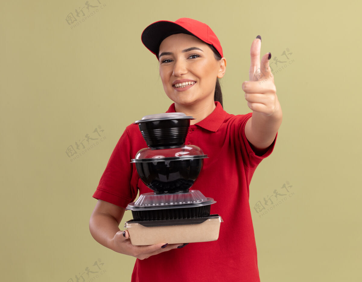 盒子身穿红色制服 戴着帽子的年轻送货员 手里拿着一叠食品包 食指指着前面 站在绿色的墙边愉快地微笑着欢呼叠着食物