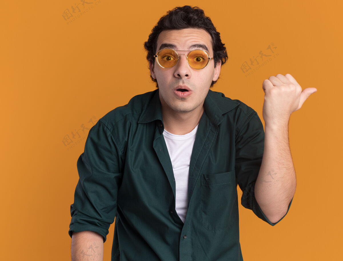 侧身穿绿衬衫戴眼镜的年轻人困惑地看着前面 用大拇指指着站在橙色墙上的一边站指点衬衫