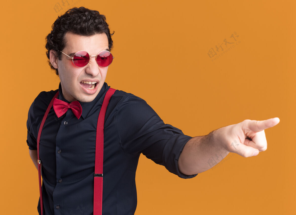 目录愤怒的时髦男人戴着蝴蝶结 戴着眼镜和吊带 用食指指着站在橙色墙上的一边眼镜时尚弓