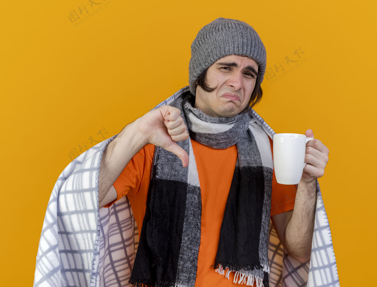 放下一个戴着冬天帽子 围巾裹着格子布 拿着一杯茶 拇指朝下 被隔离在橙色背景下的未出狱的年轻人包裹茶围巾