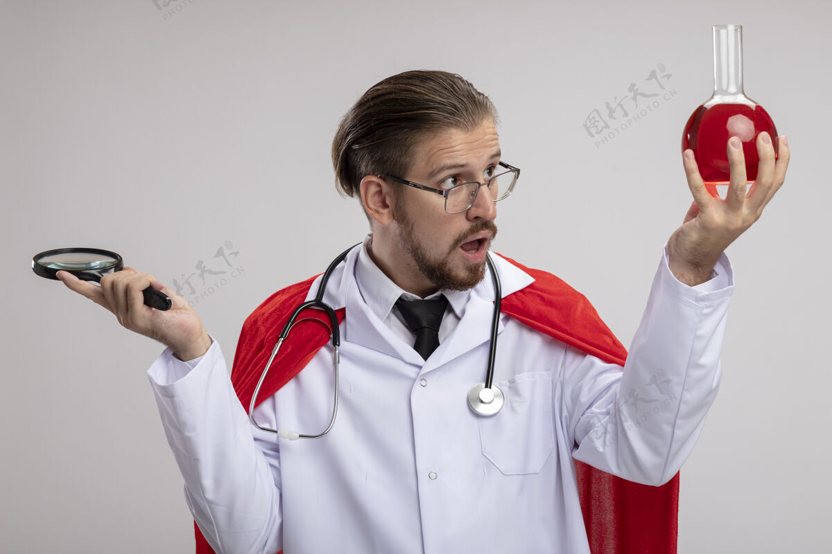 听诊器一个年轻的超级英雄穿着医用长袍 戴着听诊器和眼镜 拿着放大镜 看着他手上装满红色液体的化学玻璃瓶 被隔离在白色背景上填补家伙惊喜