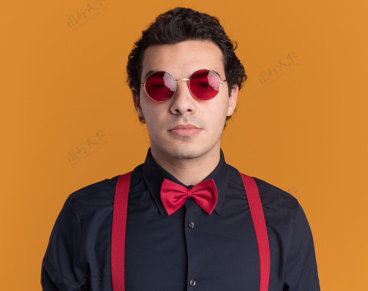 自信戴着眼镜 系着吊带 打着领结的时髦男人站在橙色的墙上 神情严肃自信地看着前方领带眼镜站立