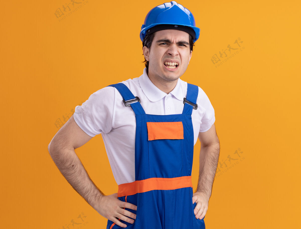 安全年轻的建筑工人穿着建筑制服 戴着安全帽 站在橙色的墙上 怒气冲冲地看着前面建筑失望头盔