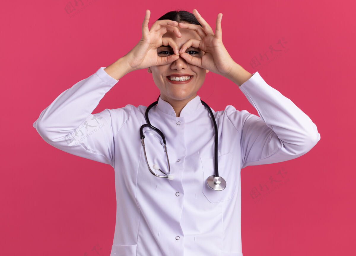 使身穿医用外套的年轻女医生手持听诊器 用手指注视着前方 做着双目手势 站在粉红色的墙上开心地微笑着望远镜微笑手势
