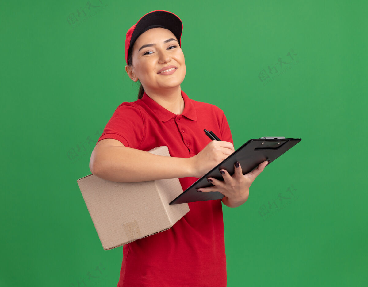 微笑身穿红色制服 头戴鸭舌帽的年轻女送货员拿着一个纸板箱 上面写着写字板 面带微笑 自信地站在绿色的墙上看着前面制服帽子女人