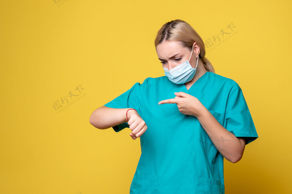 成人身穿医用衬衫 戴着消毒口罩的年轻女医生的正视图看着她在黄色墙上的手腕手腕面具Lookign
