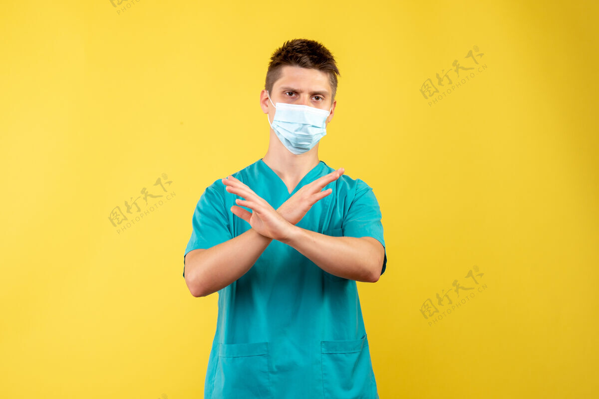 前面黄色墙壁上穿着医疗服和无菌口罩的男医生的正视图健康人西装