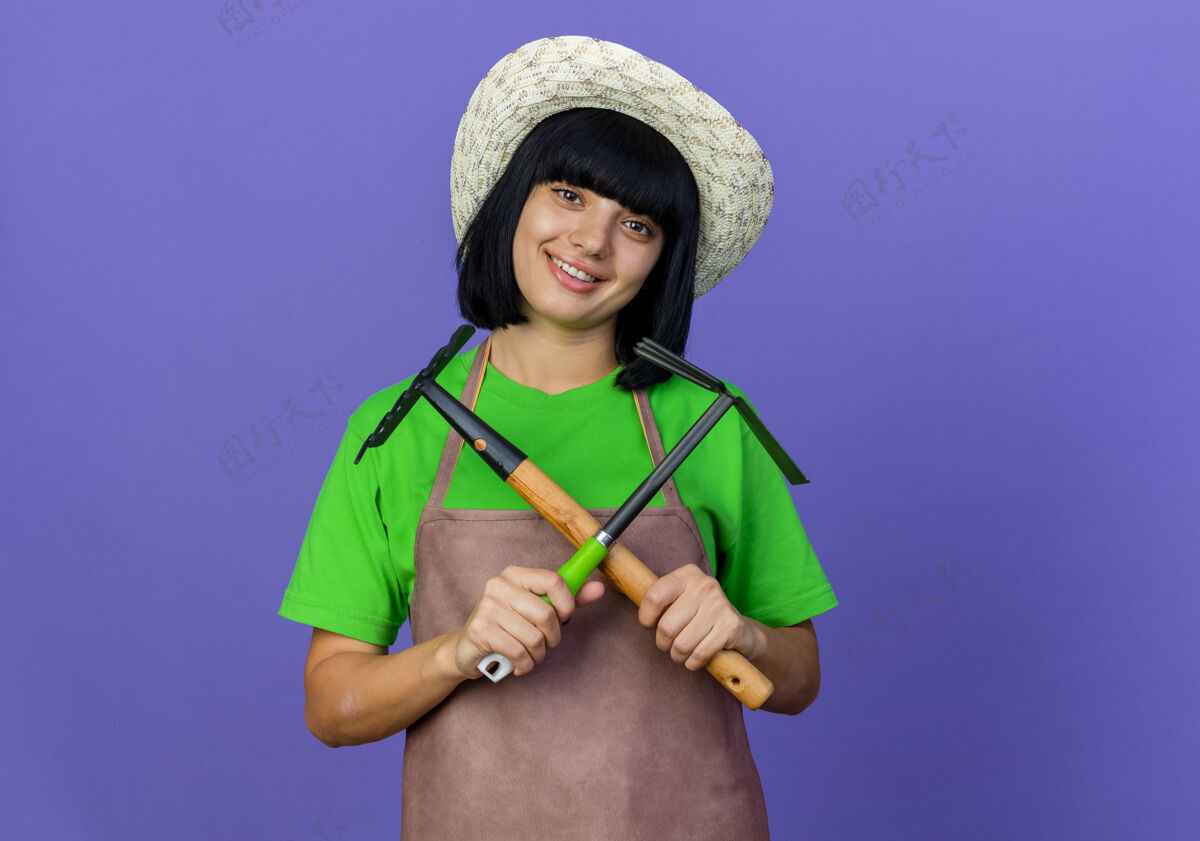 制服微笑的年轻女园丁穿着制服 戴着园艺帽 手持交叉耙和锄头耙复制紫色年轻
