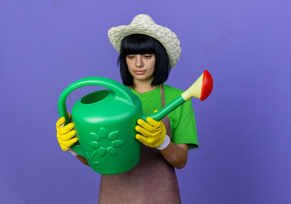 手套自信的年轻女园丁穿着制服 戴着园艺帽和手套 拿着并看着浇水罐紫色穿空间