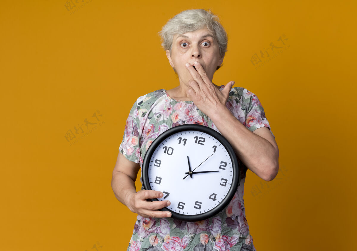 拿着震惊的老妇人把手放在嘴巴上 拿着橙色墙上的钟手老人桔子
