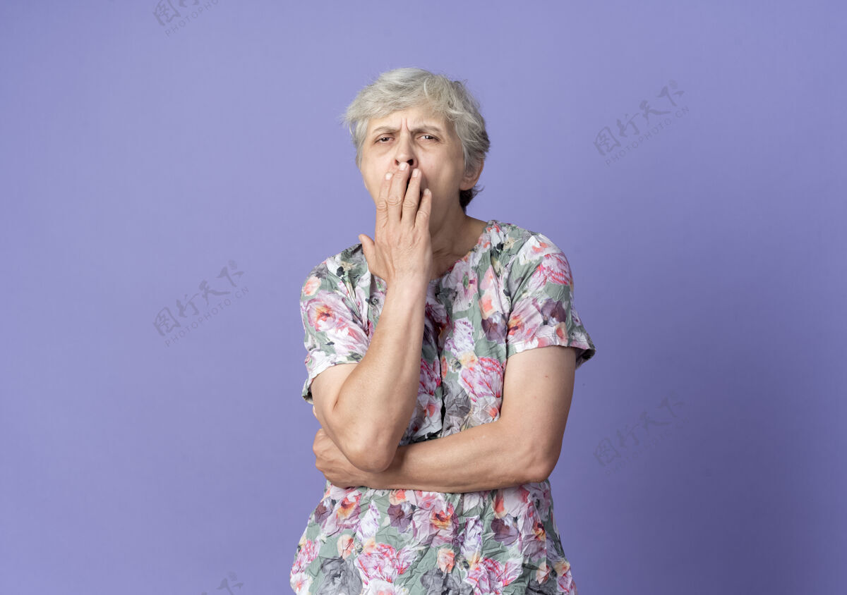 困困倦的老妇人打着哈欠 把嘴孤立地放在紫色的墙上抱嘴巴哈欠