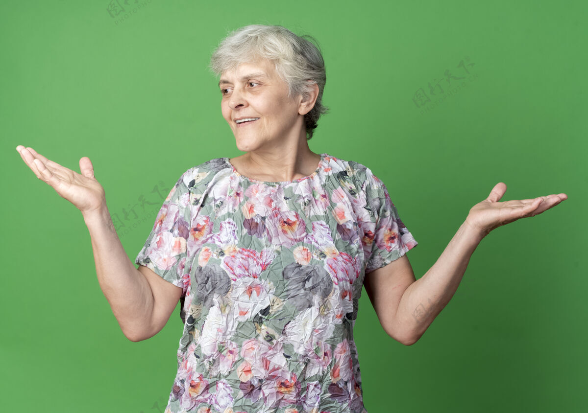 老微笑的老妇人举起双手站在绿色的墙上 看着孤立的一面绿女人举起来