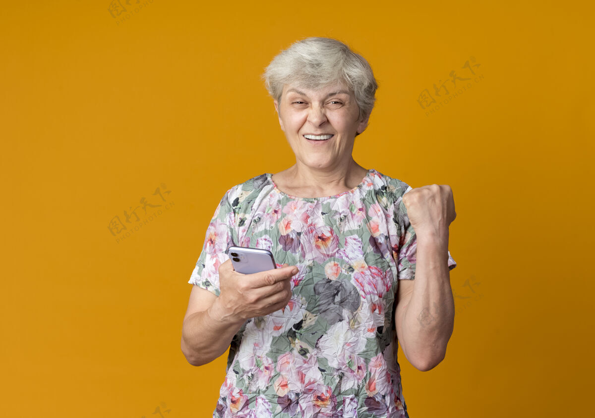 电话微笑的老妇人把握紧电话的拳头孤立在橙色的墙上微笑女人保持