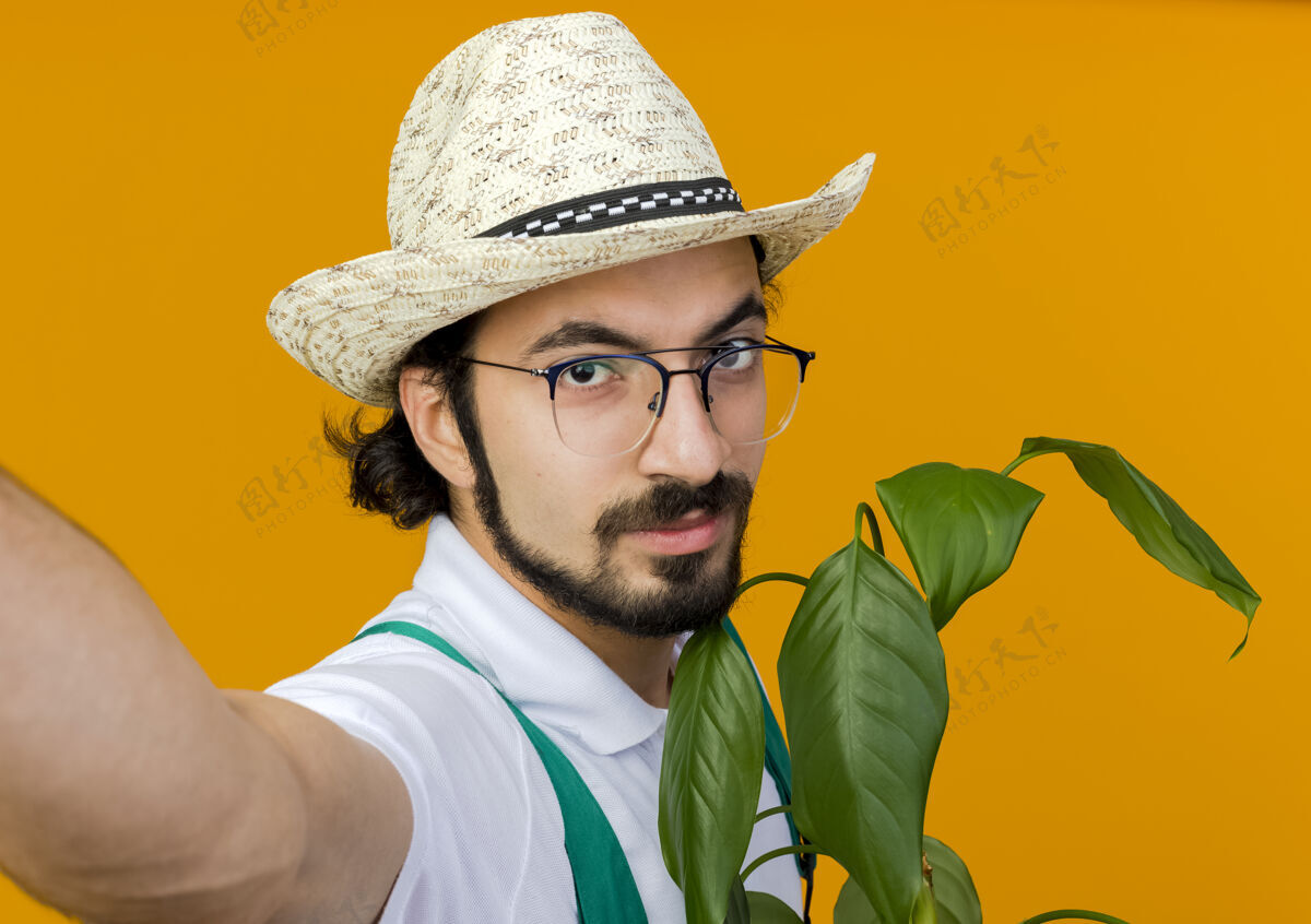 眼镜自信的男园丁戴着眼镜 戴着园艺帽 拿着植物 假装拿着相机穿着园艺空间