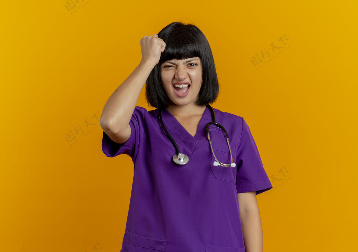 头恼怒的年轻黑发女医生穿着制服用听诊器把拳头放在孤立的橙色背景和复印空间的头上年轻拳头女性