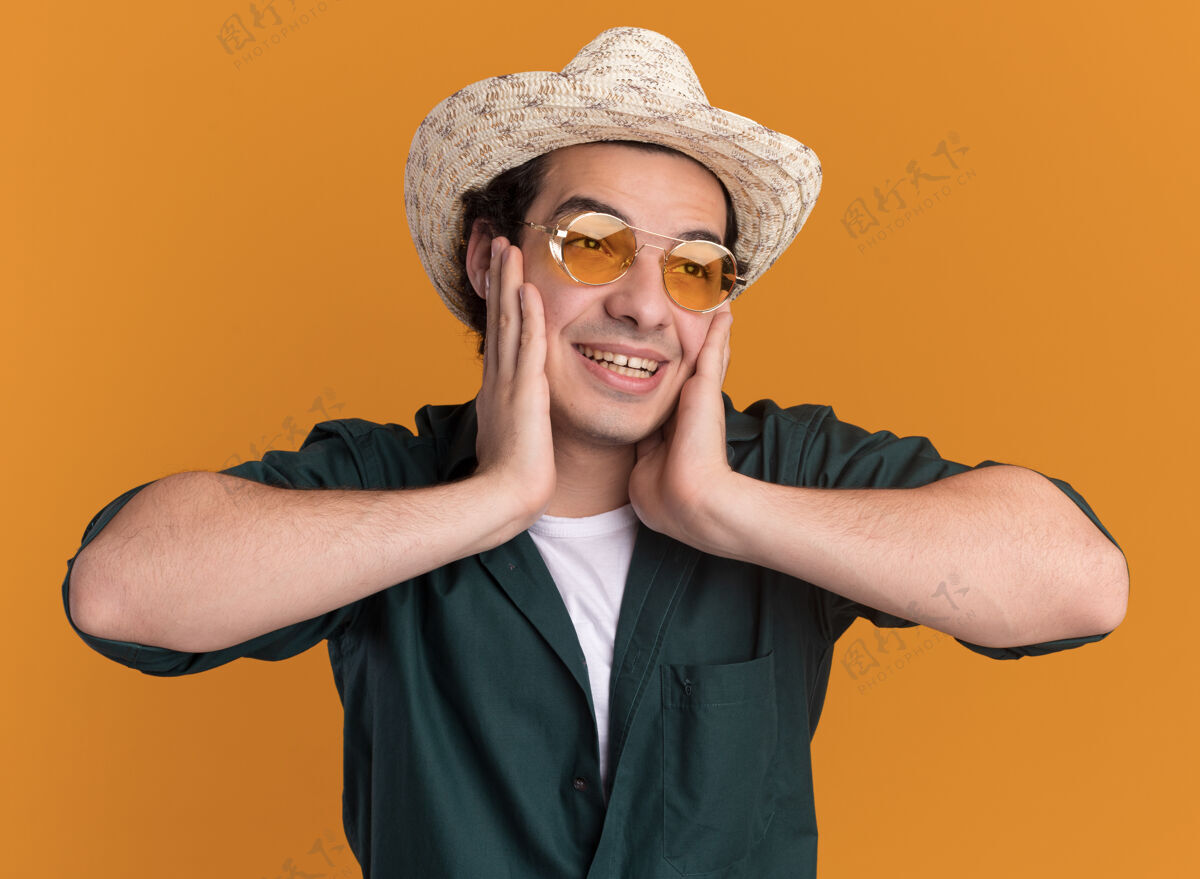 戴着穿着绿色衬衫 戴着夏天帽子 戴着眼镜 脸上带着微笑的快乐的年轻人站在橙色的墙上夏天放在一边帽子