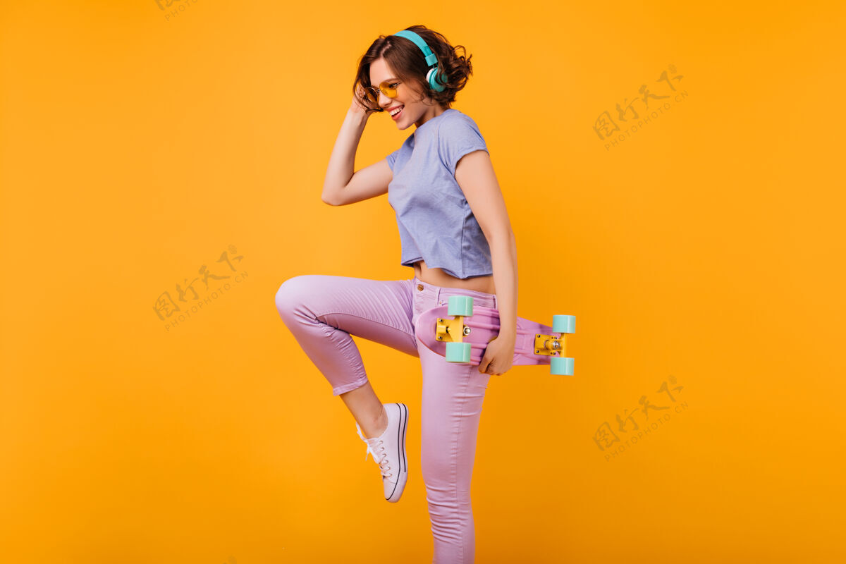 多彩苗条迷人的女孩 戴着蓝色耳机 微笑着跳舞身材匀称的女模特 跳着跳板 脸上洋溢着幸福的表情微笑玩滑板