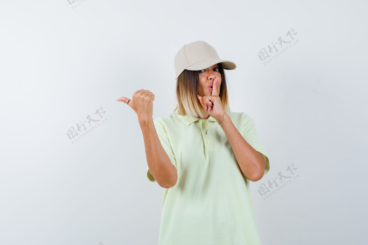 时尚年轻女士展示沉默的手势 同时用拇指指着左边 穿着t恤 戴着帽子 看起来很自信 正面视图成人前面左边