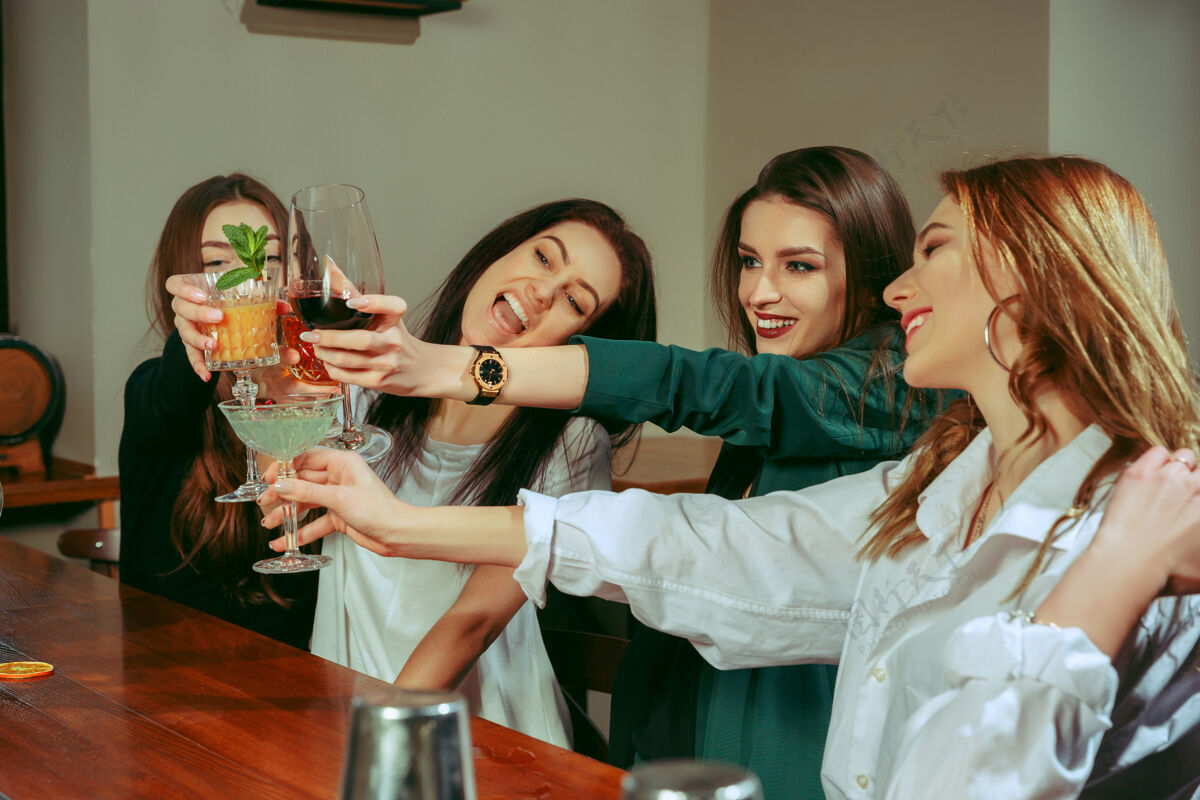 一起女朋友们在酒吧喝酒 她们坐在一张摆着鸡尾酒的木桌旁 她们在碰杯酒杯子团体