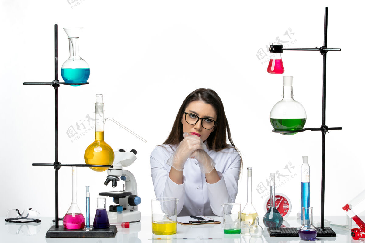 坐着前视图穿着白色医疗服的女化学家坐在白色办公桌上 拿着不同的解决方案科学大流行病毒冠状病毒实验室西装容器大流行