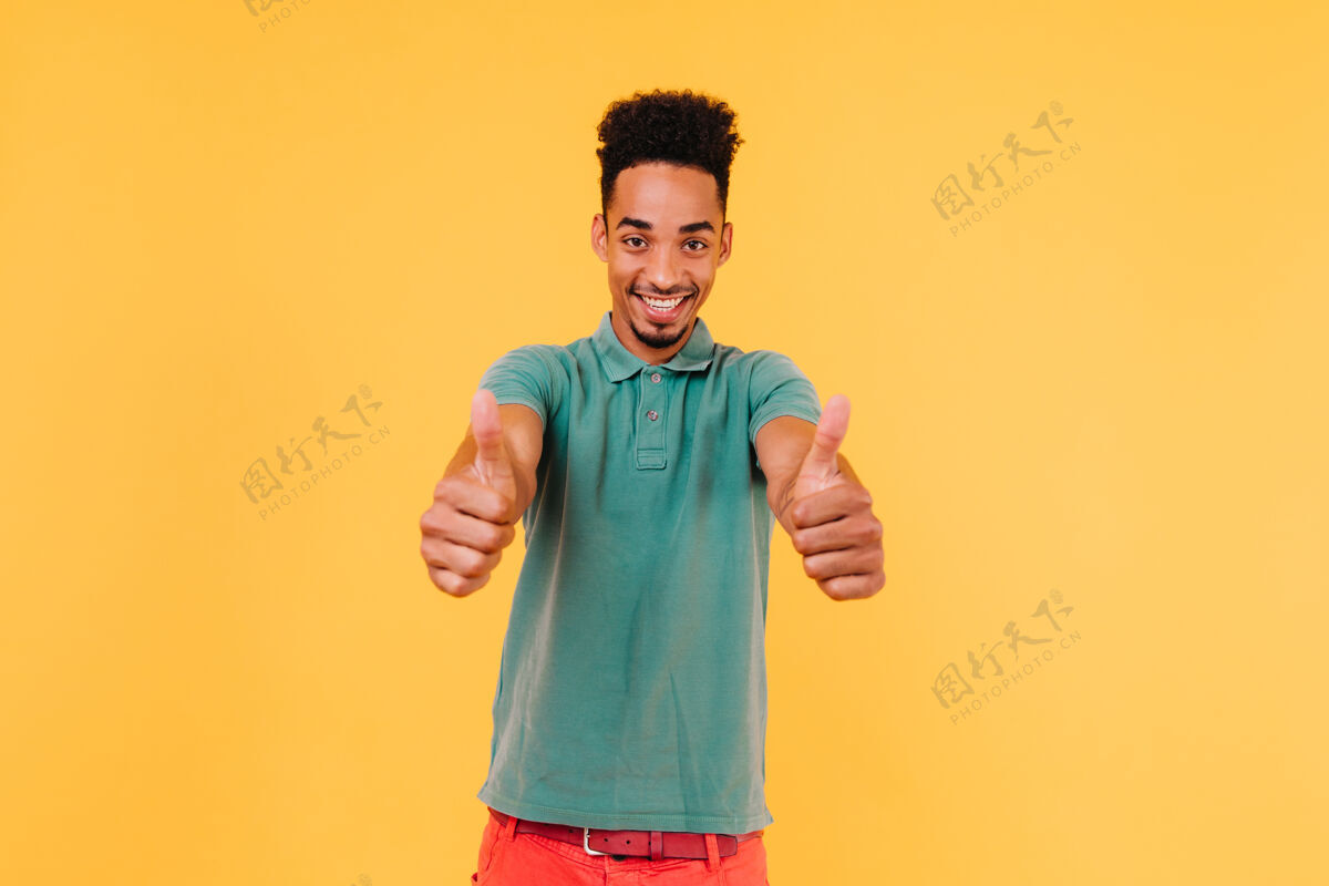 放松穿着休闲绿色t恤的无忧无虑的黑人微笑着情绪激动的非洲男人竖起大拇指摆姿势的室内照片卷发快乐明亮