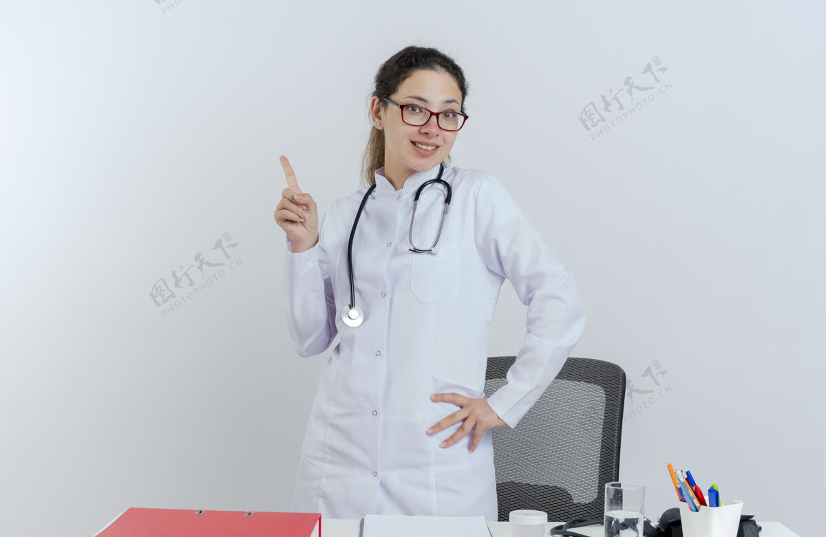 眼镜面带微笑的年轻女医生 穿着医用长袍 戴着听诊器和眼镜 站在桌子后面 手里拿着医疗工具 手放在腰上 抬起手指桌子微笑举着