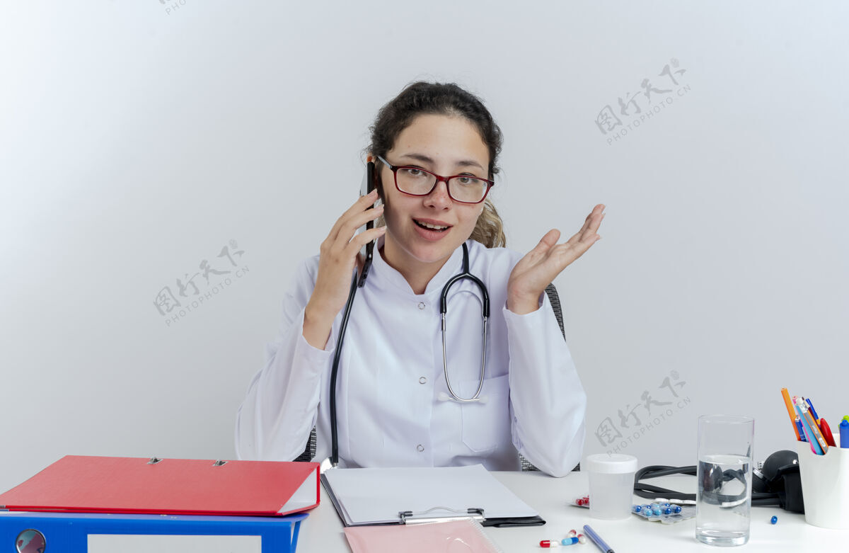 人年轻的女医生穿着医用长袍 戴着听诊器 戴着眼镜 坐在办公桌旁 拿着医疗工具 讲着电话 看上去空手孤立印象表演桌子