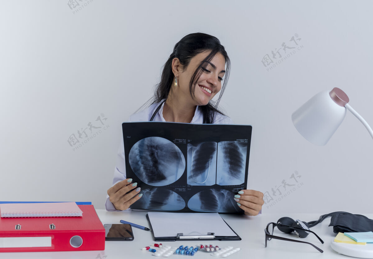 工具面带微笑的年轻女医生穿着医用长袍和听诊器坐在办公桌旁 手持医疗工具 看着x光片年轻人成人穿着