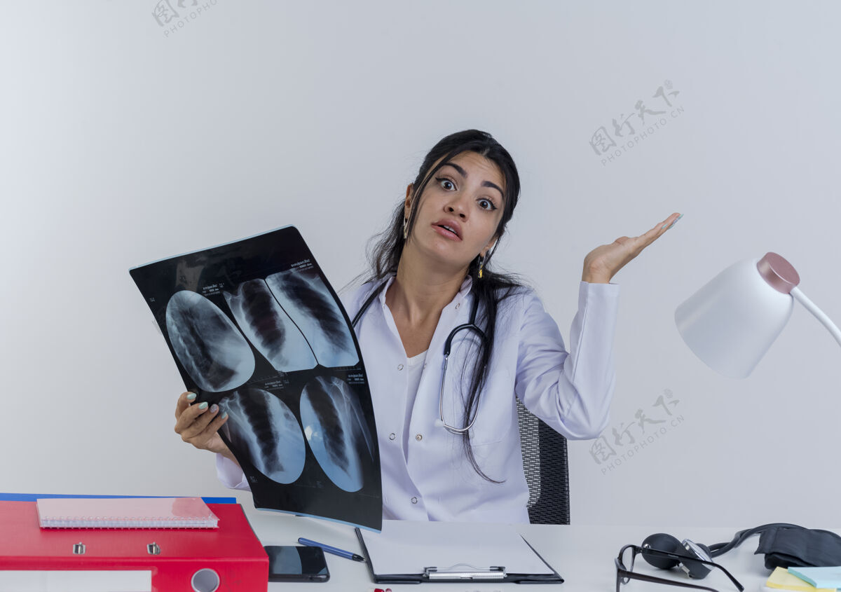 抱着困惑的年轻女医生穿着医用长袍 听诊器坐在办公桌旁 手持医疗工具 拿着x光片 看样子空手孤立年轻女人射击