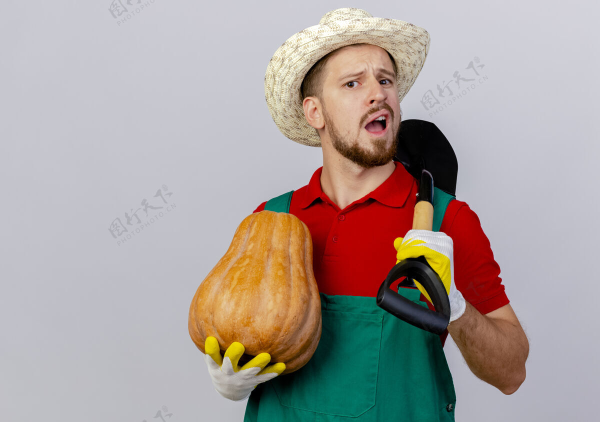 秋天年轻英俊的斯拉夫园丁穿着制服 戴着园艺手套和帽子 手里拿着南瓜和铲子 看上去与世隔绝年轻人花园南瓜