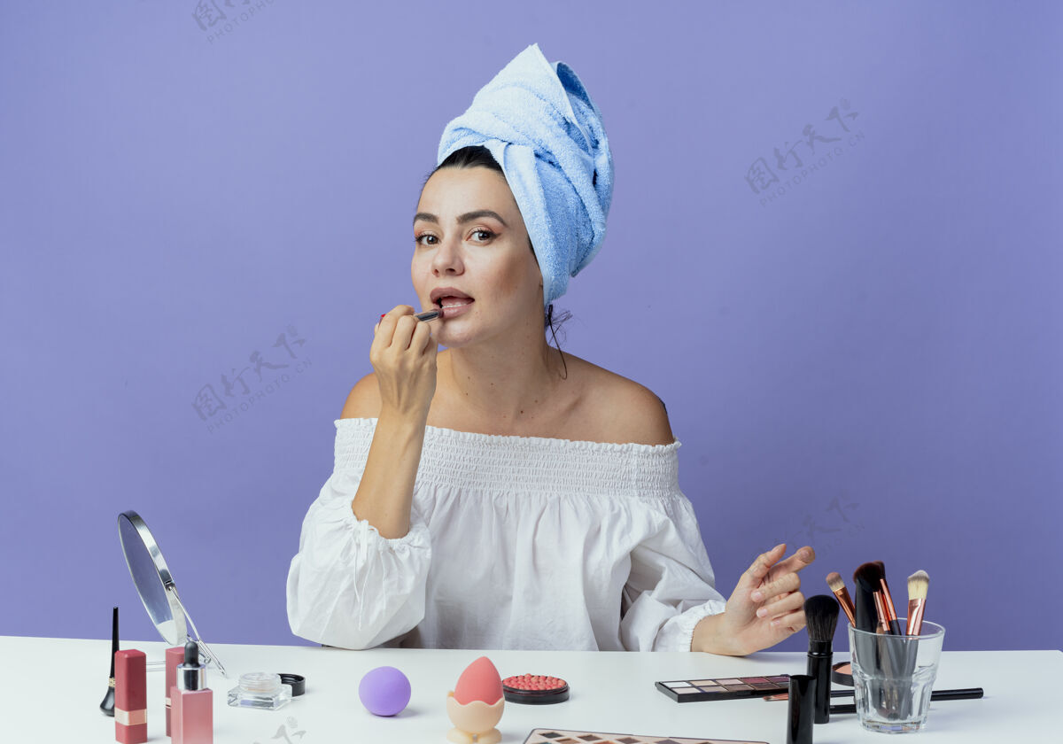 保持自信漂亮的女孩裹着发巾坐在桌子上 拿着化妆工具 拿着唇膏 看着紫墙上的孤零零女人桌子口红