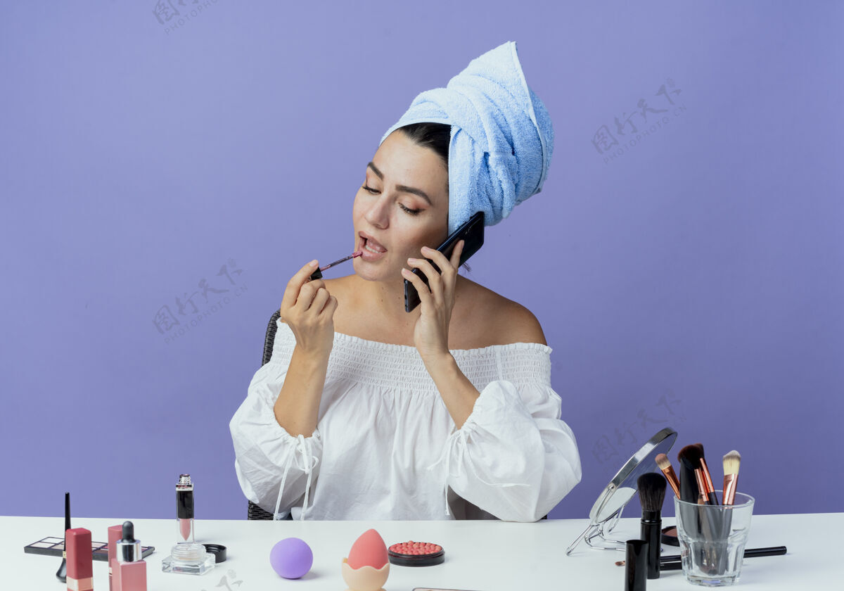 紫色自信漂亮的女孩裹着发巾坐在桌子上 用化妆工具拿着唇彩 用手机在紫壁上隔着电话交谈包裹嘴唇光泽