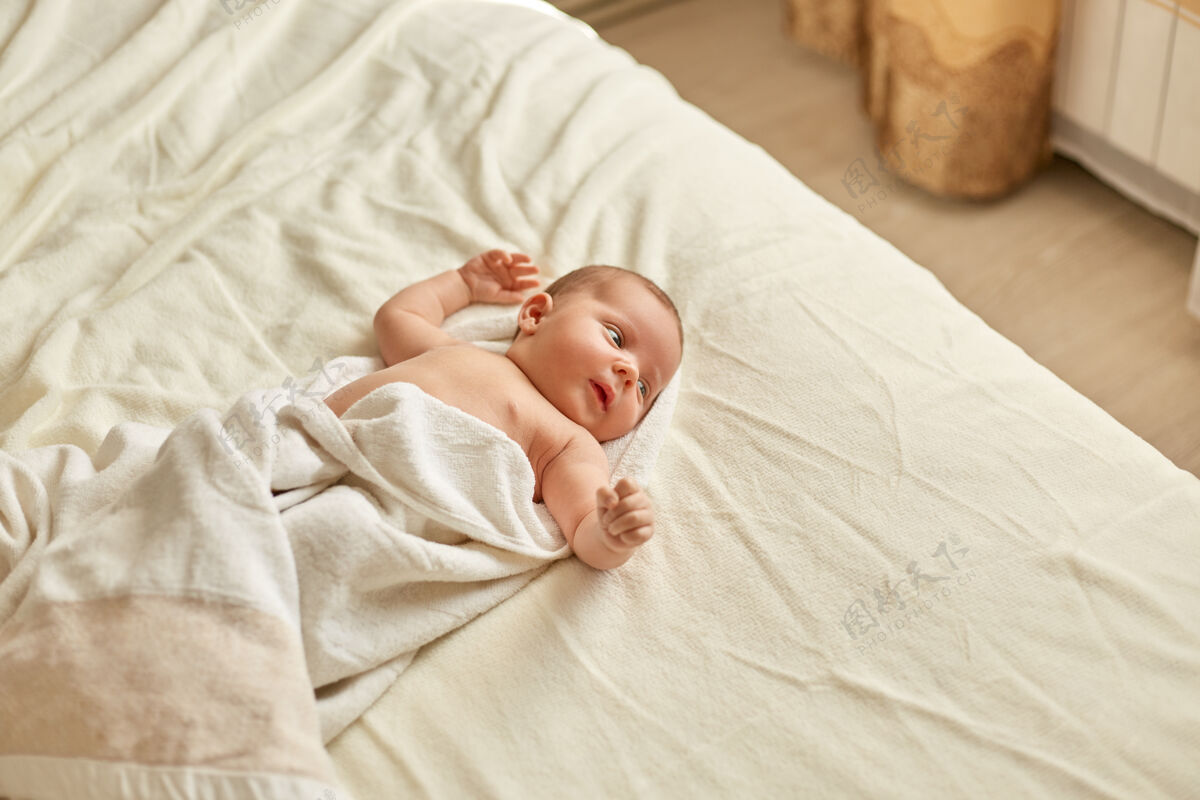 好奇可爱的宝宝洗澡后裹着毛巾躺在床上 白色格子布 望着别处举手 宝宝学习外面的东西 室内的小男孩或小女孩有趣婴儿孩子